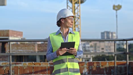 Portrait-shot-of-the-young-Caucasian-pretty-blond-woman-constructor-or-foreman-in-hardhat-and-goggles-with-tablet-device-in-hands-turning-face-to-the-camera-and-smiling-cheerfully-at-the-building-site.